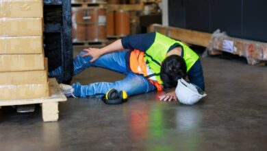 A warehouse worker lies on the ground after a fall, holding their head down and clutching at their knee.