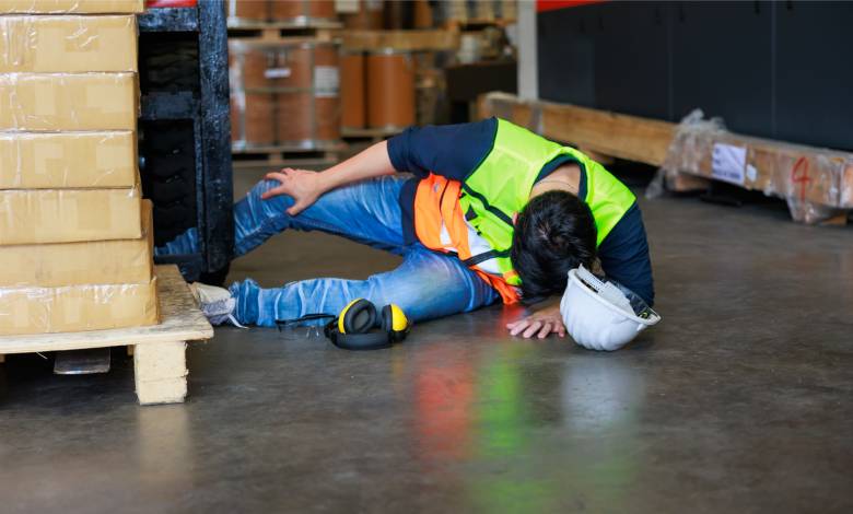 A warehouse worker lies on the ground after a fall, holding their head down and clutching at their knee.
