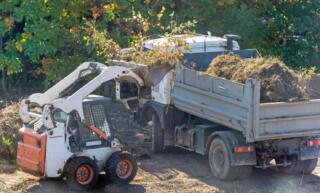 A skid steer operator uses the bucket attachment to put sticks and grass into a dump truck during land clearing work.
