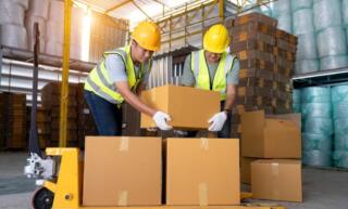 Two male warehouse workers are working together to lift sealed cardboard boxes onto a pallet for shipment.