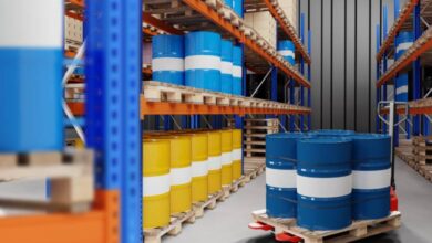 Blue and yellow metal barrels are organized on warehouse racks. Between two rows of racks, a pallet jack holds four barrels.