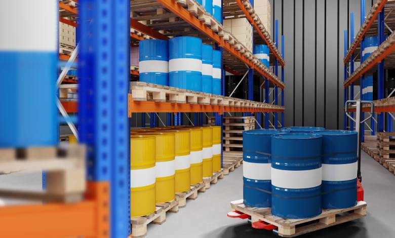 Blue and yellow metal barrels are organized on warehouse racks. Between two rows of racks, a pallet jack holds four barrels.