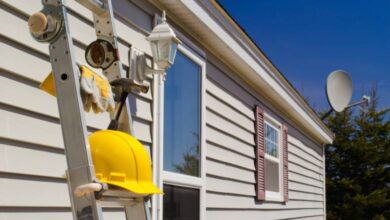 A ladder, holding a yellow hard hat and a pair of work gloves, leans against the side of a house with new siding.