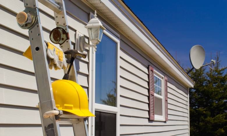 A ladder, holding a yellow hard hat and a pair of work gloves, leans against the side of a house with new siding.