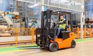 A man in a high visibility vest operating an orange and black forklift in an open area on a manufacturing floor.