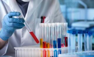 A lab employee wears a white lab coat and examines several glass tubes filled with colorful liquid.