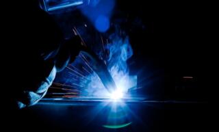 A worker in a face shield and thick gloves using a plasma welding torch. A blue aura, smoke, and sparks fly from the contact point.