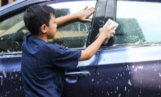 A young boy holding a white sponge in his right hand, using it to wash a blue car. The car is covered with soap and water.