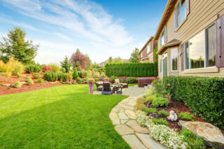 A grand backyard landscape with a small outdoor living area. Bushes line the house and tall hedges line the side fence.