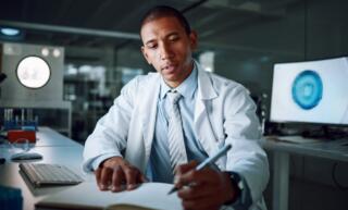 A lab manager sitting at a desk in his laboratory at night, writing in a book with a pen. He wears a white lab coat.