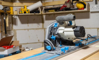 An electric track saw sitting on a piece of wood at a worktable. Other woodworking tools are in the background.