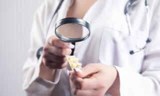 A woman wearing a doctor's lab coat and stethoscope examines a package of yellow pills with a magnifying glass.