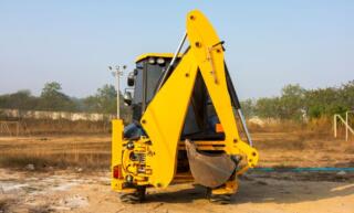 A skid steer with an excavator attachment parked in a field. There are metal structures and a line of trees in the background.