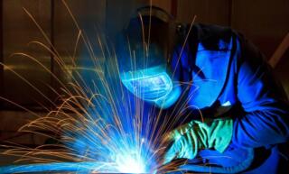 An industrial employee wearing a welding helmet and heavy welding clothes uses a welder with sparks flying.