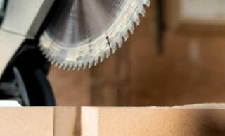 A close-up view shows a silver, circular saw blade hovering above a piece piece of sandy-colored wood.