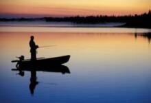 A silhouette of a man fishing at sunrise in a small boat on a lake. The sky and water are blue, orange, and purple.
