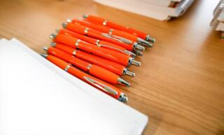 A bunch of identical orange pens on a wooden desk, lying between a stack of paper and rows of stacked binders.