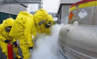 Three individuals in yellow hazmat suits, one holding a fire extinguisher, conduct a drill simulating a deadly gas leak.