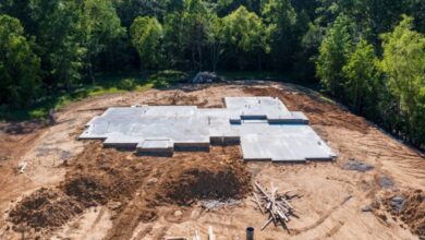 A home construction site features a freshly-poured concrete slab foundation surrounded by cleared dirt and a tree line.