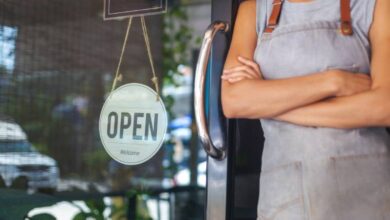 A woman stands in the doorway of a small business. She wears an apron and has her arms folded. A small sign says "open" on the door.