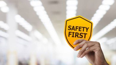 A hand holding a yellow sign that reads "safety first" in black, capitalized letters. There are rows of lights behind the sign.