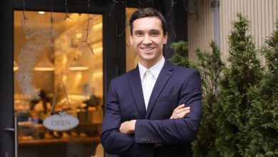 A man stands in front of a business with his arms folded and smiling. An "Open" sign hangs on the business's door.