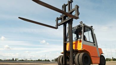 A yellow forklift with its forks lifted to the top of its mast is parked outside in an empty lot on a sunny, partly cloudy day.