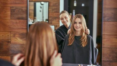 A woman sits in a black salon chair with a black cape around her body. She looks in a mirror as another woman touches her hair.