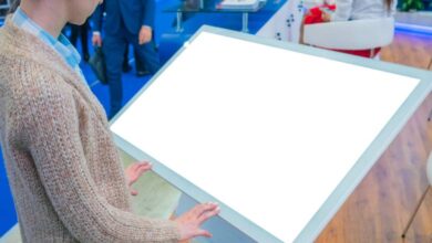 A person in a tan cardigan stands in front of a large, white touchscreen at a trade show. The screen is blank.