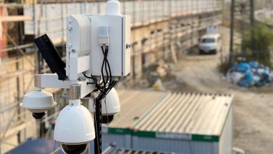Modern security cameras are mounted on top of a pole near a large, industrial construction site near storage containers.