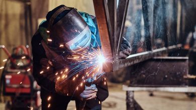 A welder in full PPE bending over to weld the joint of a metal-beam structure. Blue and orange sparks are flying.