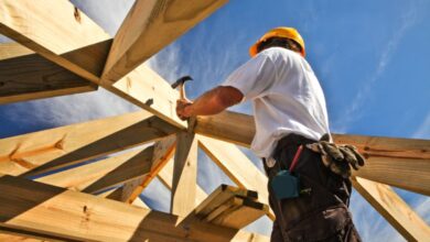 A man in a white shirt, jeans, and yellow hard hat hammers a nail into a wooden plank during construction.