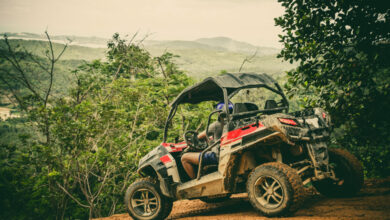A person wearing a gray shirt, blue shorts, and a blue helmet sits in the driver’s seat of a red UTV on a muddy path.