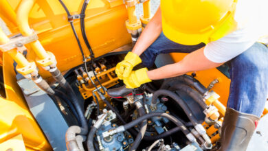 A person sitting by the engine of a construction vehicle and working on it with a wrench. They are wearing a hard hat and gloves.