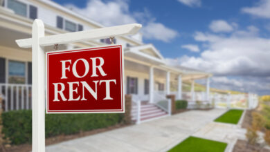A red and white sign contains the words for rent. It sits on the lawn of a white home with a large front porch.