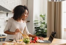 A woman standing in a kitchen mixing salad ingredients and chopping vegetables while looking at her laptop.