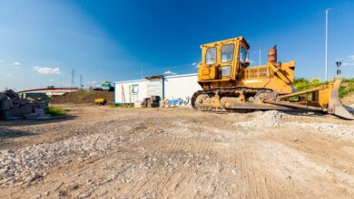 A yellow excavator is parked in front of a white building at a construction site near piles of rocks.