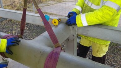 Two workers wearing yellow reflective jackets and blue gloves as they wrap purple rigging slings around a pipe.