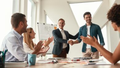 Two men stand at the head of a large table, shaking hands with each other. Two women and a man sit at the table, clapping.