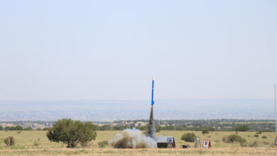A small white and blue model rocket is surrounded by dark smoke as it launches from a spot in the middle of a field.