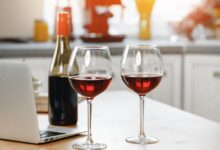 Two glasses of red wine, a half-full wine bottle, and a silver laptop sit on a kitchen island.