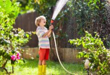 A young child smiling as they hold a hose by a garden sprayer attachment and watch the water spray out.