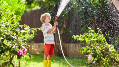 A young child smiling as they hold a hose by a garden sprayer attachment and watch the water spray out.
