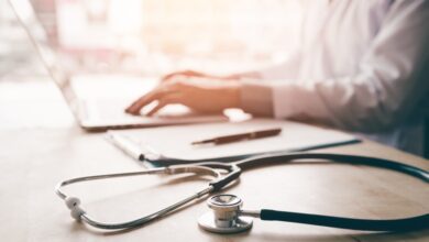 An individual in a white coat working on a laptop with a stethoscope, clipboard, and pen on the desk.