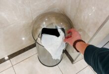 A person's hand tosses a piece of paper into a stainless steel trash can that sits in the corner of a tiled bathroom.