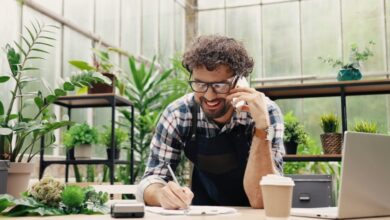 A smiling man leans on a counter, holding a cell phone up to his ear and writing on paper. Potted plants fill the background.