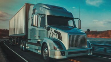 A gray semi truck is pulling a white trailer down a curvy road next to a river. Trees line the other side of the river.