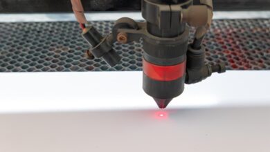 A close-up of a laser marking machine marking a blank white sheet over metal grates. A small red dot shines on the white surface.