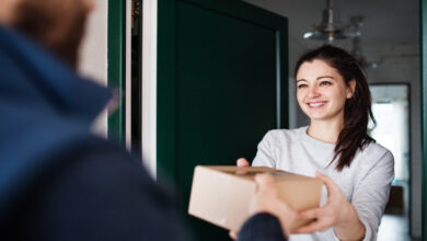 A smiling person standing in their doorway and receiving a cardboard package from a delivery person.