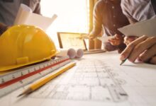 A group of people stand around a blueprint that's laid out on a table with a ruler, pencil, yellow hard hat, and a laptop.
